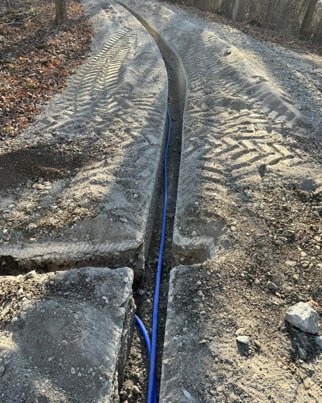 A blue pipe laid in a trench dug into a gravel road. The trench is long, and the setting appears to be outdoors.