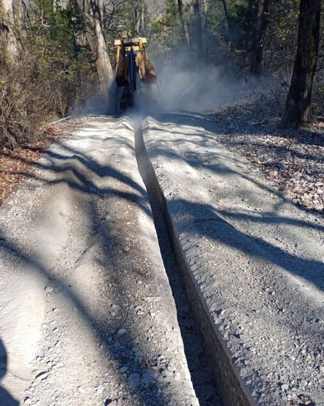 A small bulldozer cutting a deep trench into a dusty dirt road in a wooded area.