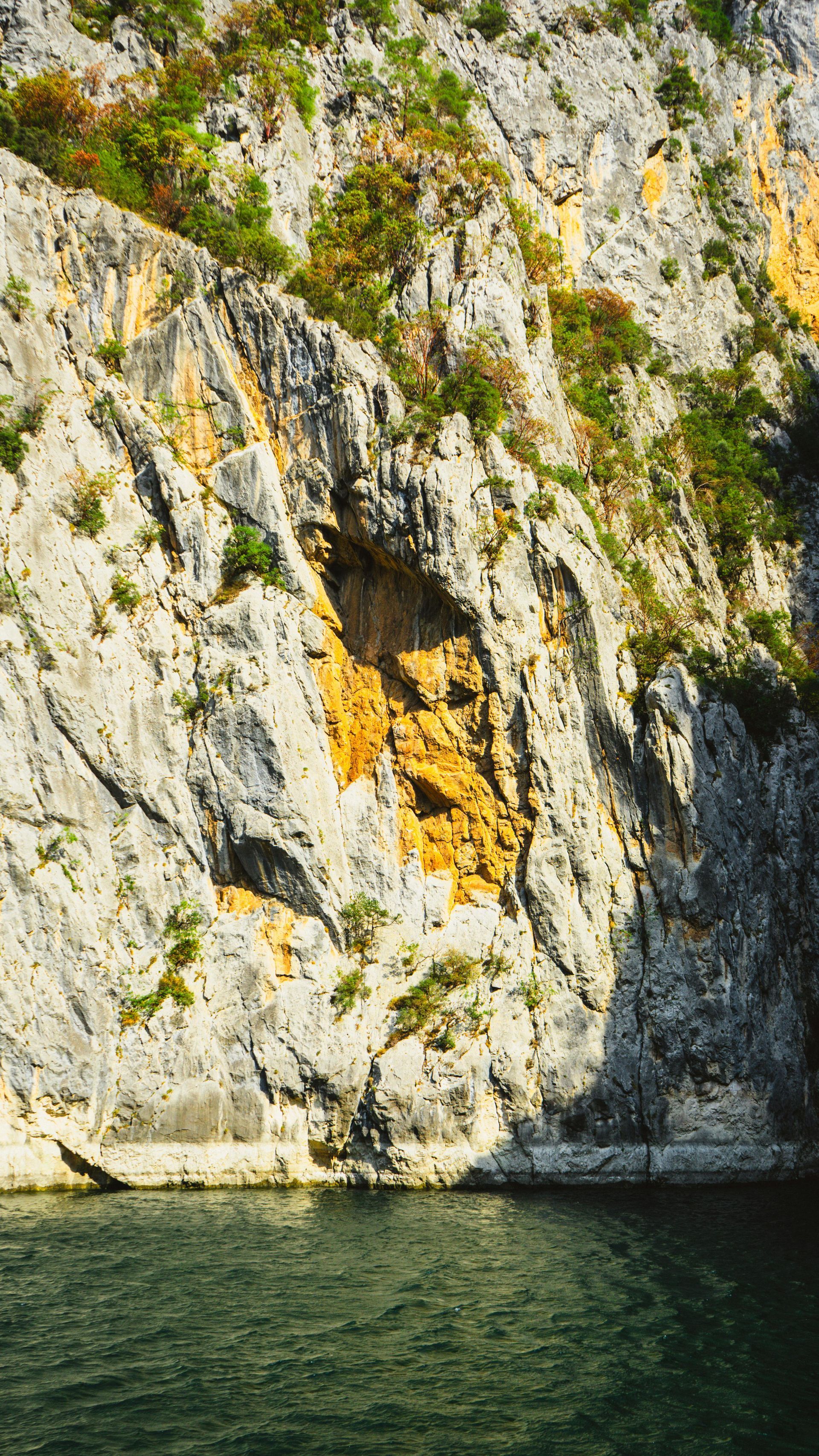 Rocky cliff face with green vegetation, beside dark water.