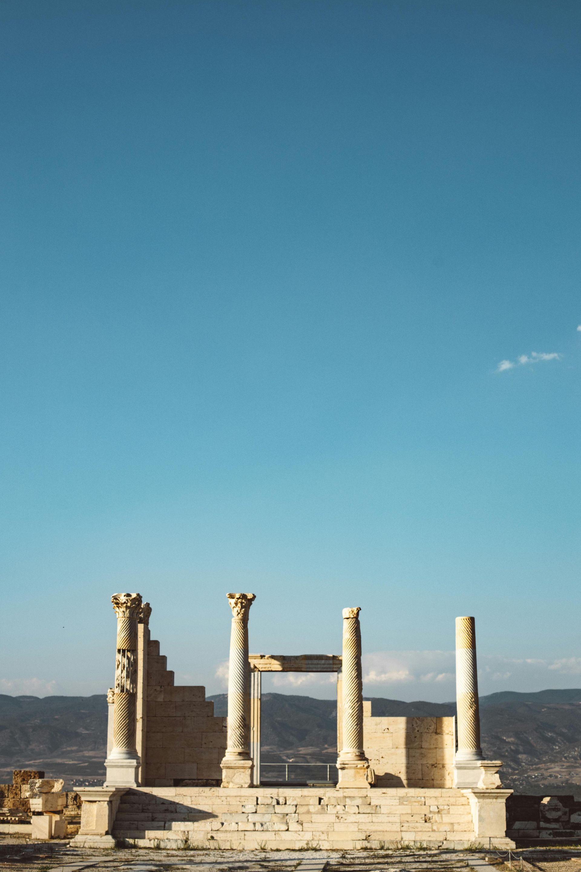 Ruins of an ancient structure with columns against a blue sky, mountains in the distance.