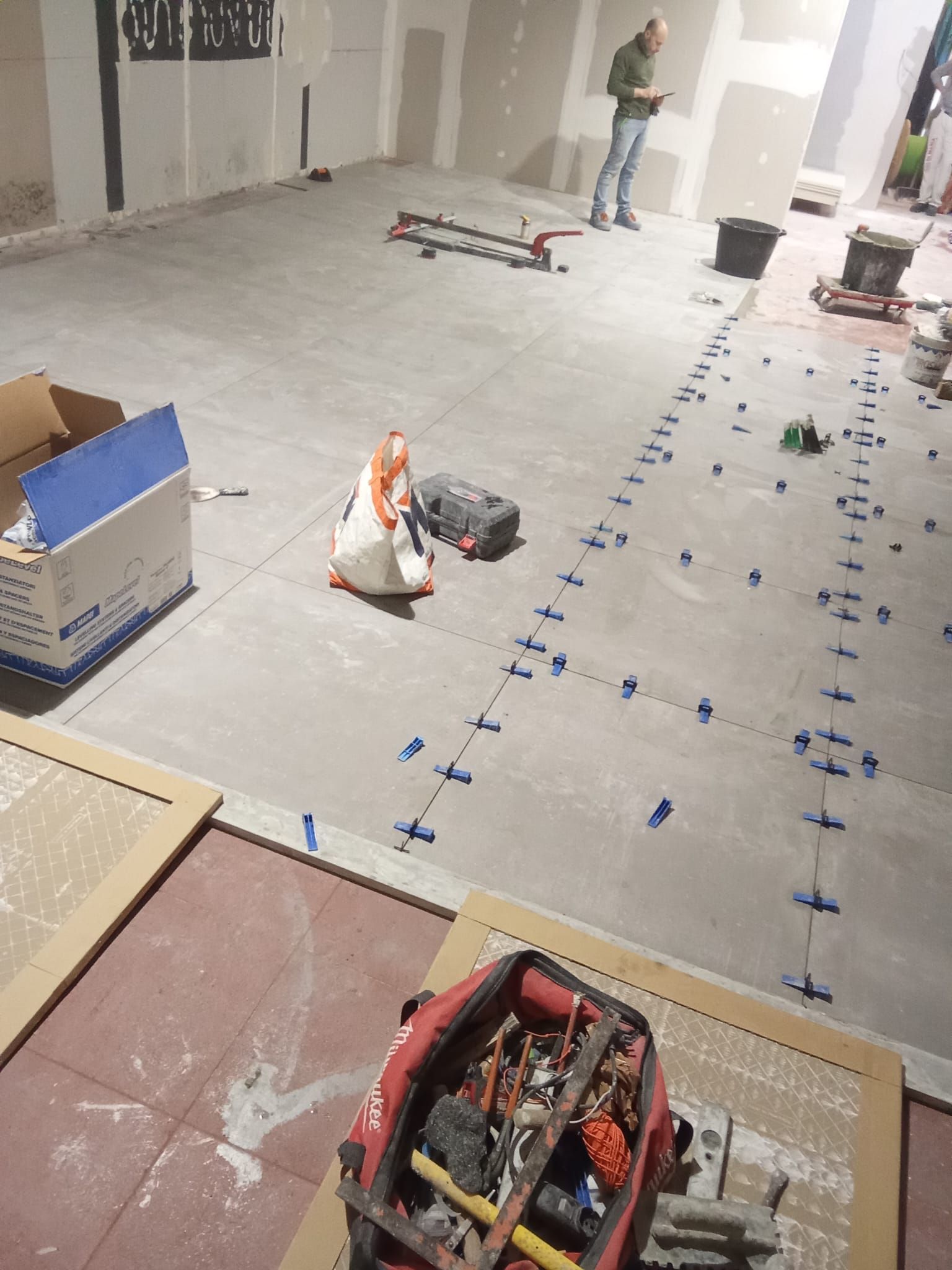 Cantiere edile con pavimento segnato, attrezzi e operai in un'ampia stanza non rifinita.