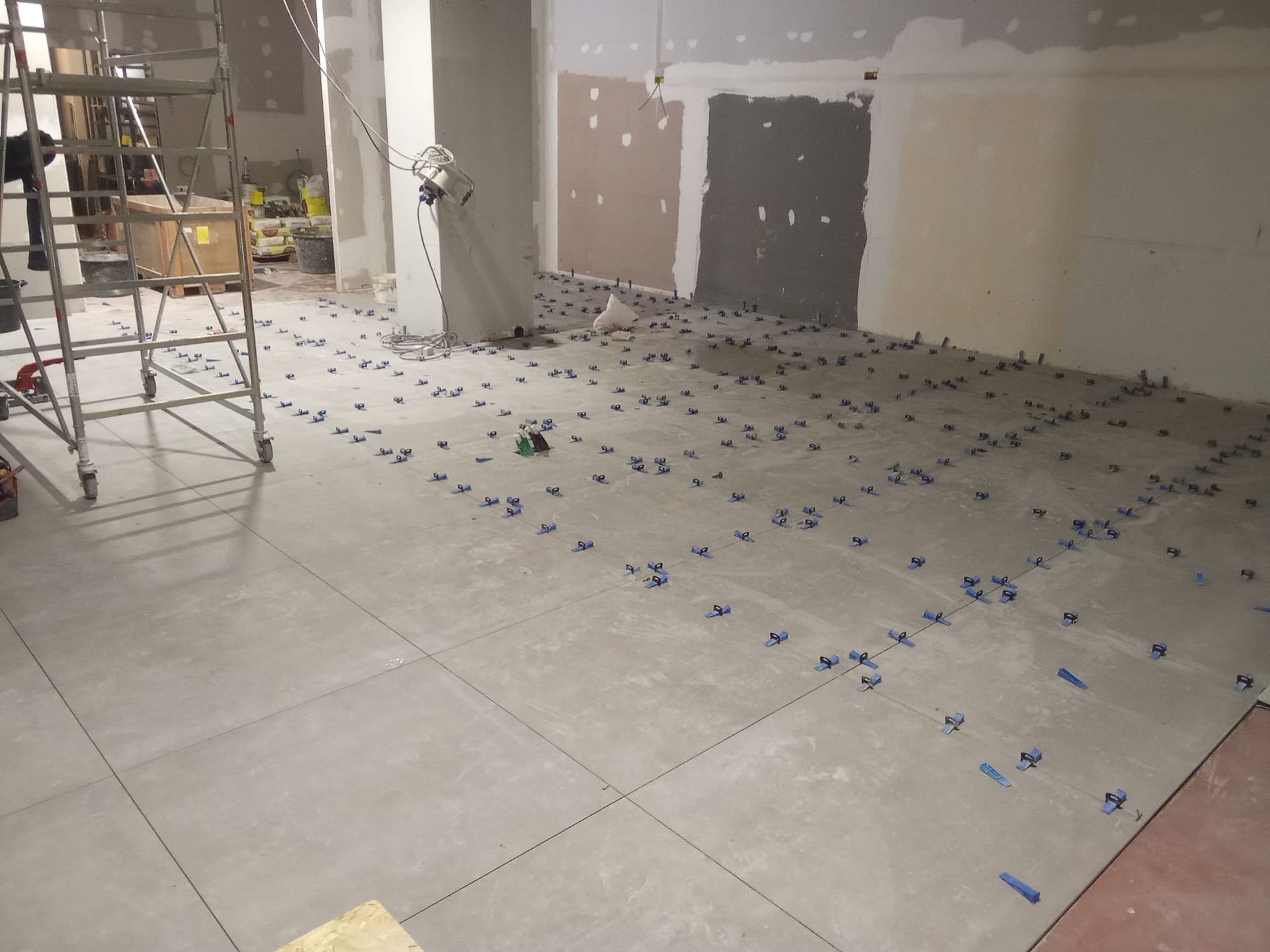 Cantiere in fase di ristrutturazione con ampia pavimentazione piastrellata, segnaletica a nastro blu, impalcature e pareti non rifinite.