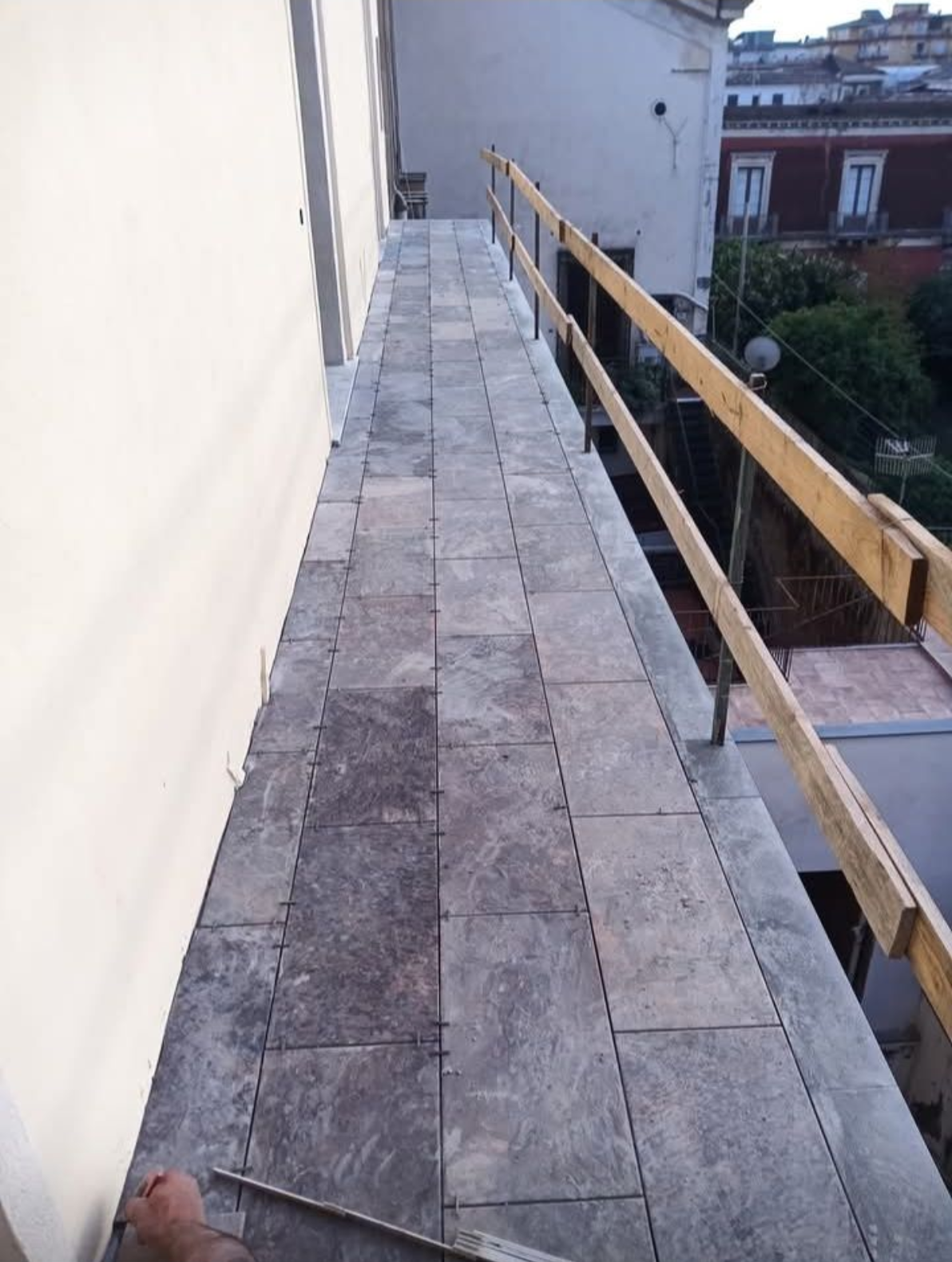 Stretta passerella sul tetto con piastrelle di pietra grigia accanto a una parete bianca e ringhiera in legno, con vista sugli edifici sottostanti.