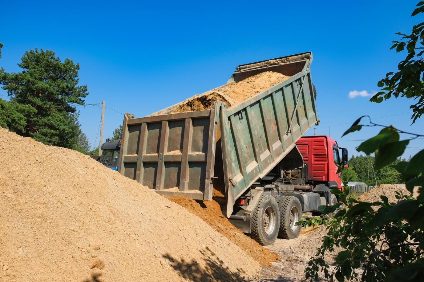 Dump truck unloading sand beside a large pile at a construction site under a blue sky