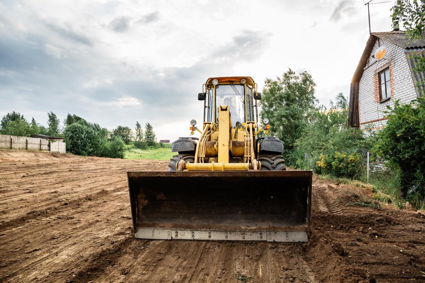 Yellow bulldozer grading dirt on a construction site beside a house and trees