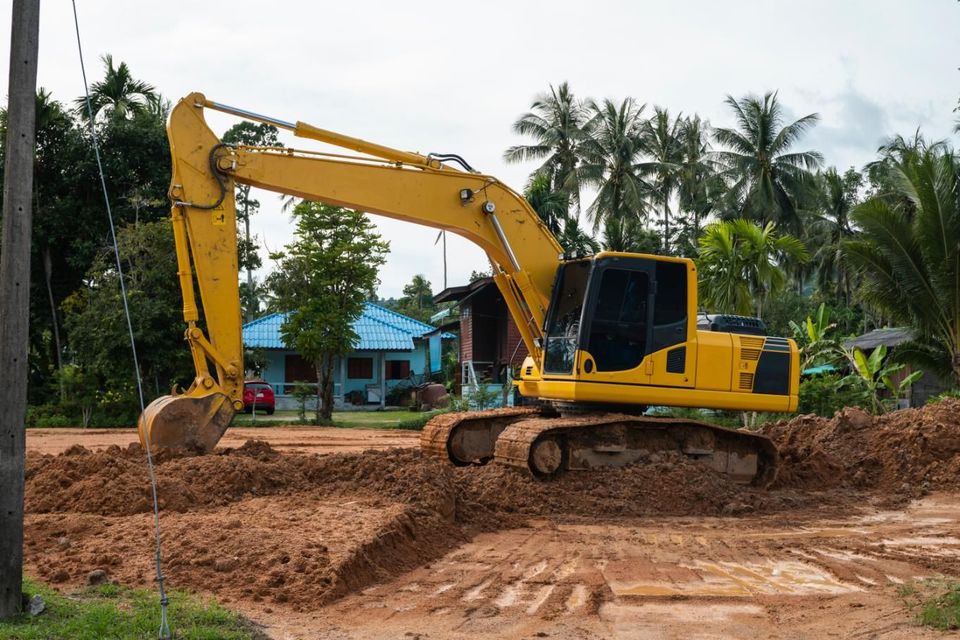 Yellow excavator digging dirt at a construction site with palm trees and a blue-roofed house in the background