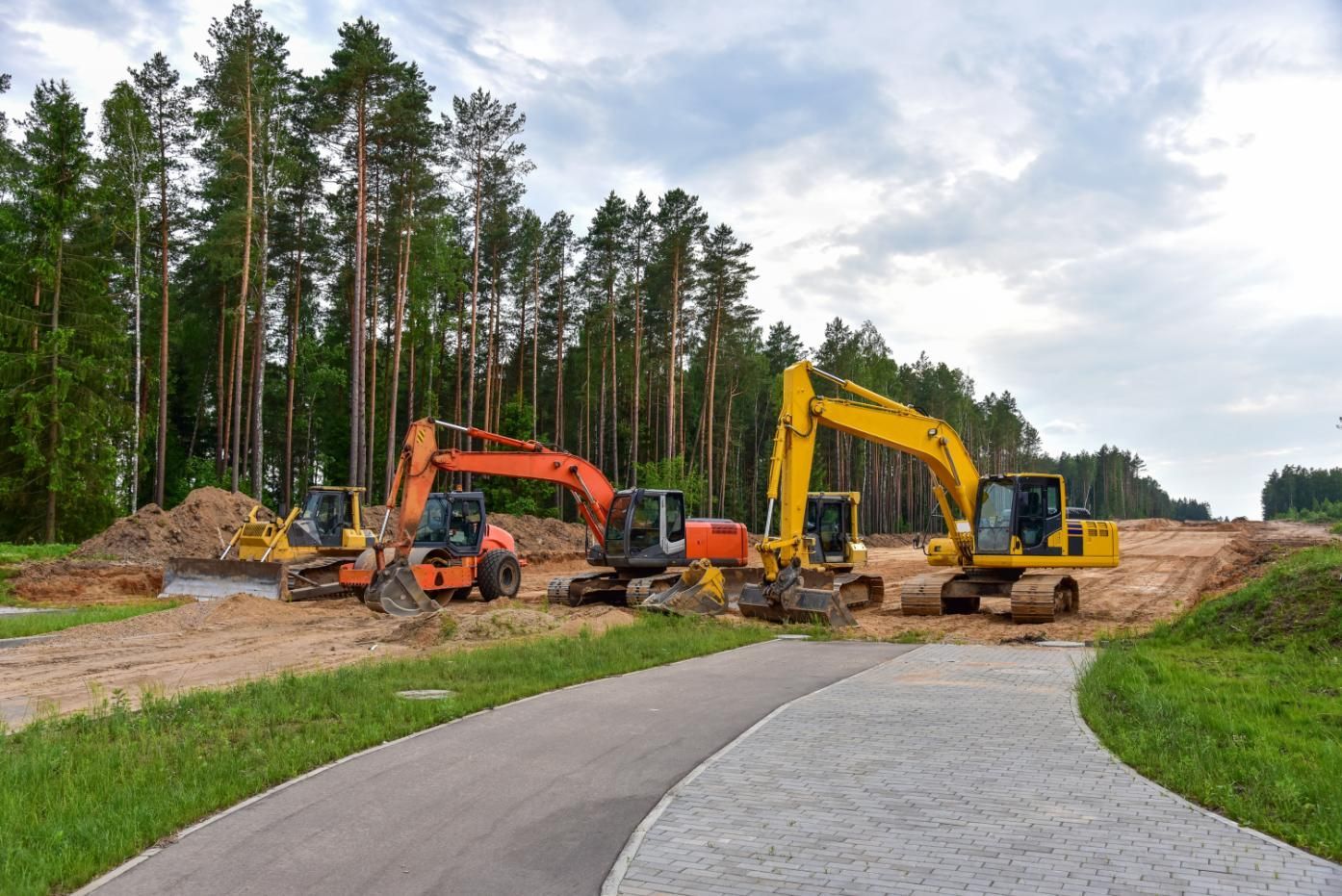 Two excavators and a dump truck on a road construction site beside a paved path and trees