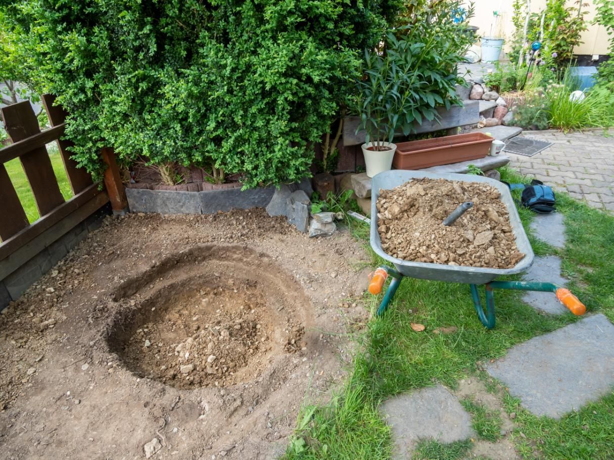 Backyard dirt holes beside a wheelbarrow full of soil near a garden fence