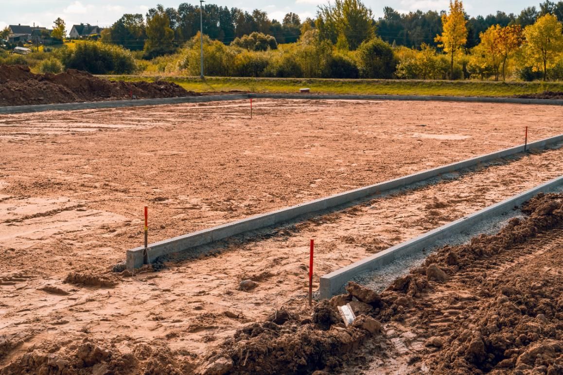 Graded construction site with two long concrete curbs set in a dirt field and trees in the background