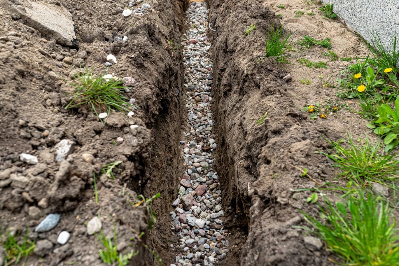 Narrow trench dug in soil with gravel bottom, surrounded by dirt and sparse grass.