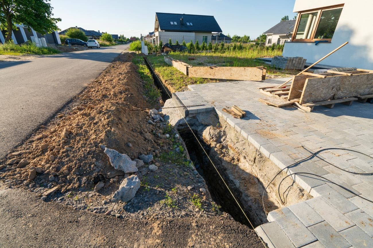 Roadside excavation beside a residential street with a trench, dirt pile, and unfinished concrete slabs