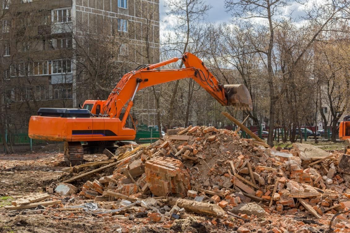 Orange excavator clearing rubble at a demolition site with apartment buildings in the background