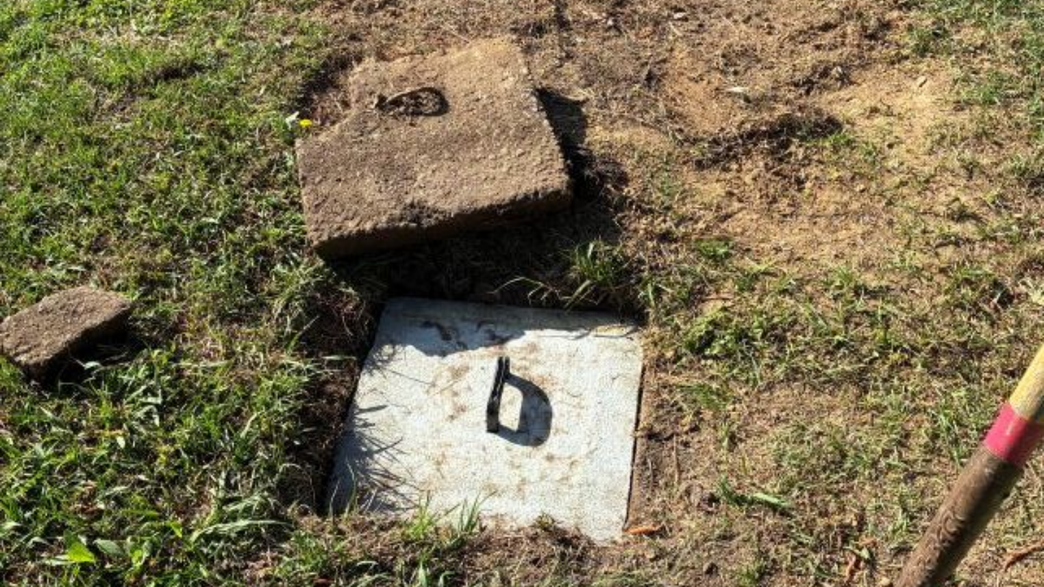Open utility access box in patchy grass beside a pink-handled tool