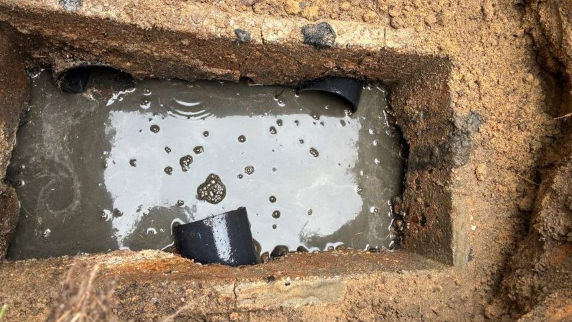 Rectangular muddy trench filled with cloudy water and a black pipe segment