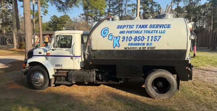White-and-blue septic service truck parked in a wooded yard, with company name and phone number on the tank.