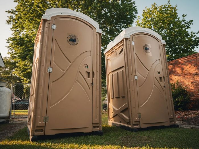 Two tan portable toilets standing on grass under trees outdoors