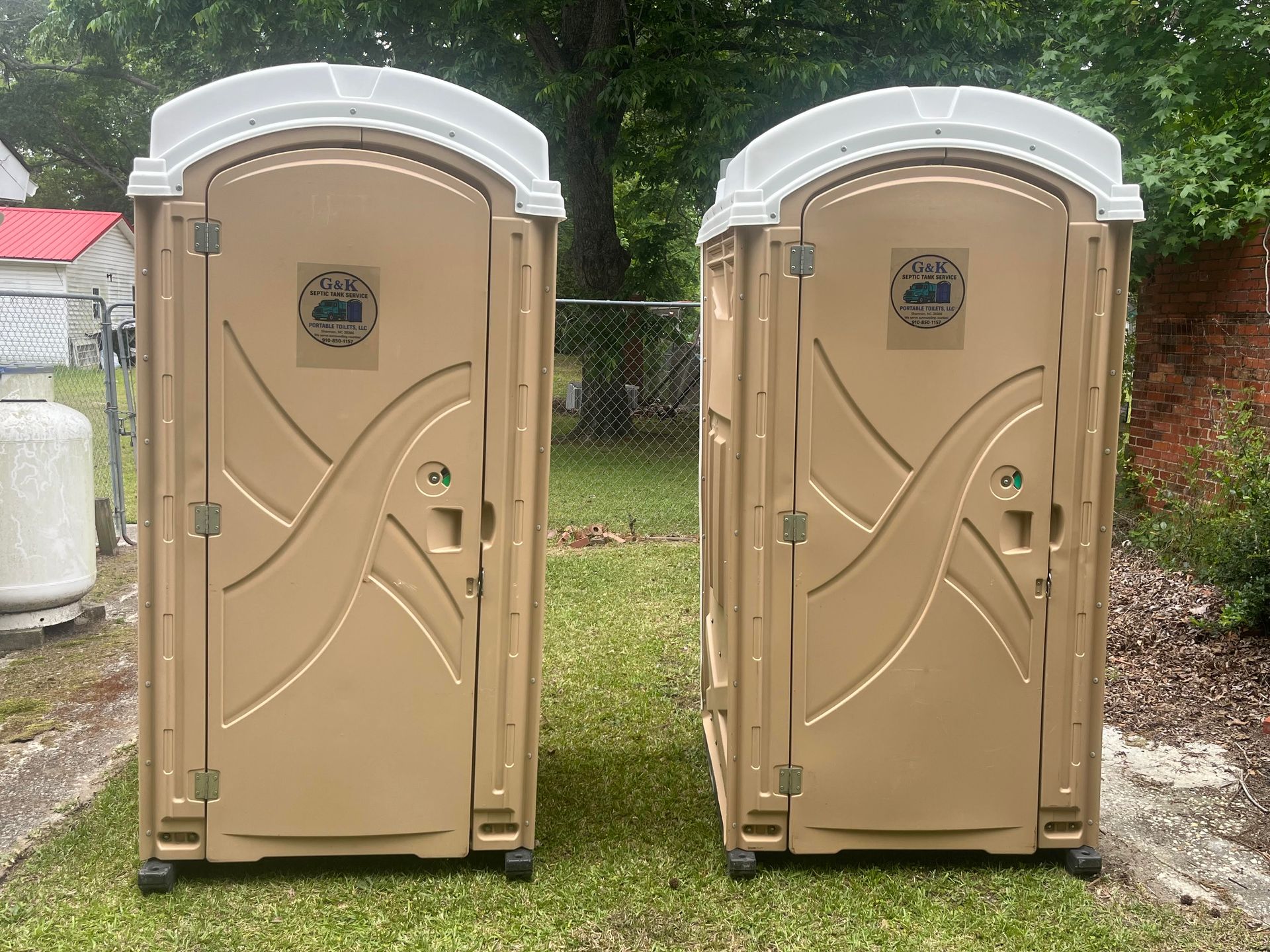 Two tan portable toilets with white roofs standing side by side outdoors.