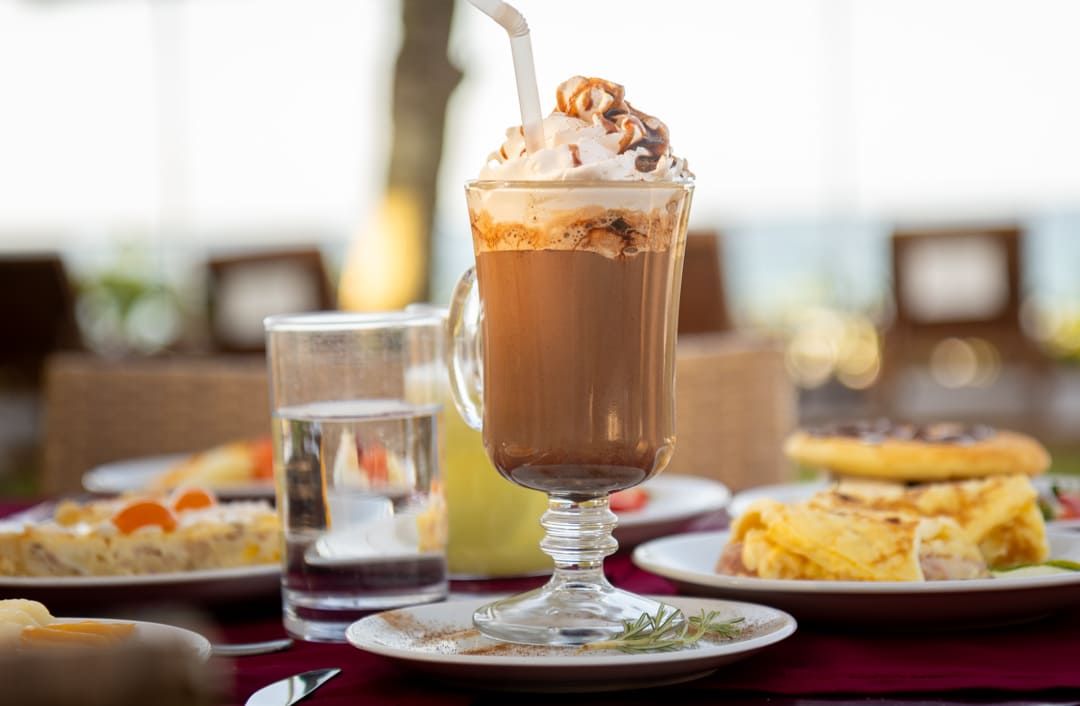 Um copo de café com chantilly por cima está sobre uma mesa com pratos de comida.