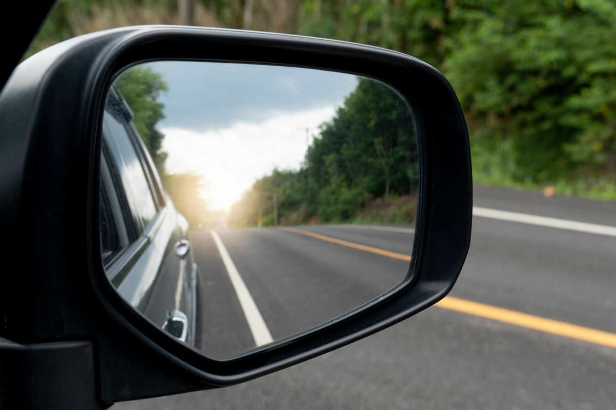 Side view mirror showing road ahead; a car's reflection on the left with trees and sky in the background.