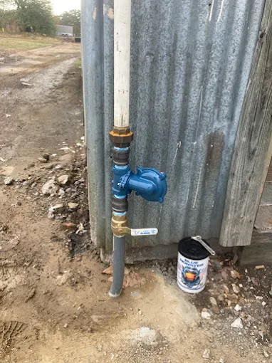Blue water valve on a vertical pipe beside a corrugated metal wall, with a dirt path and paint can nearby