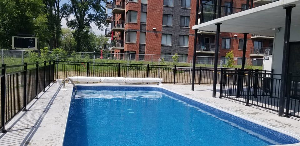 Une grande piscine est entourée d'une clôture et d'un bâtiment en arrière-plan.