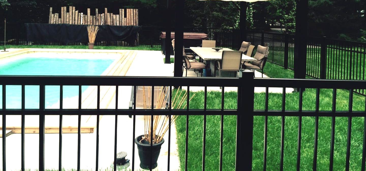 Un jardin avec piscine, terrasse et pelouse verte, visible derrière une clôture noire.