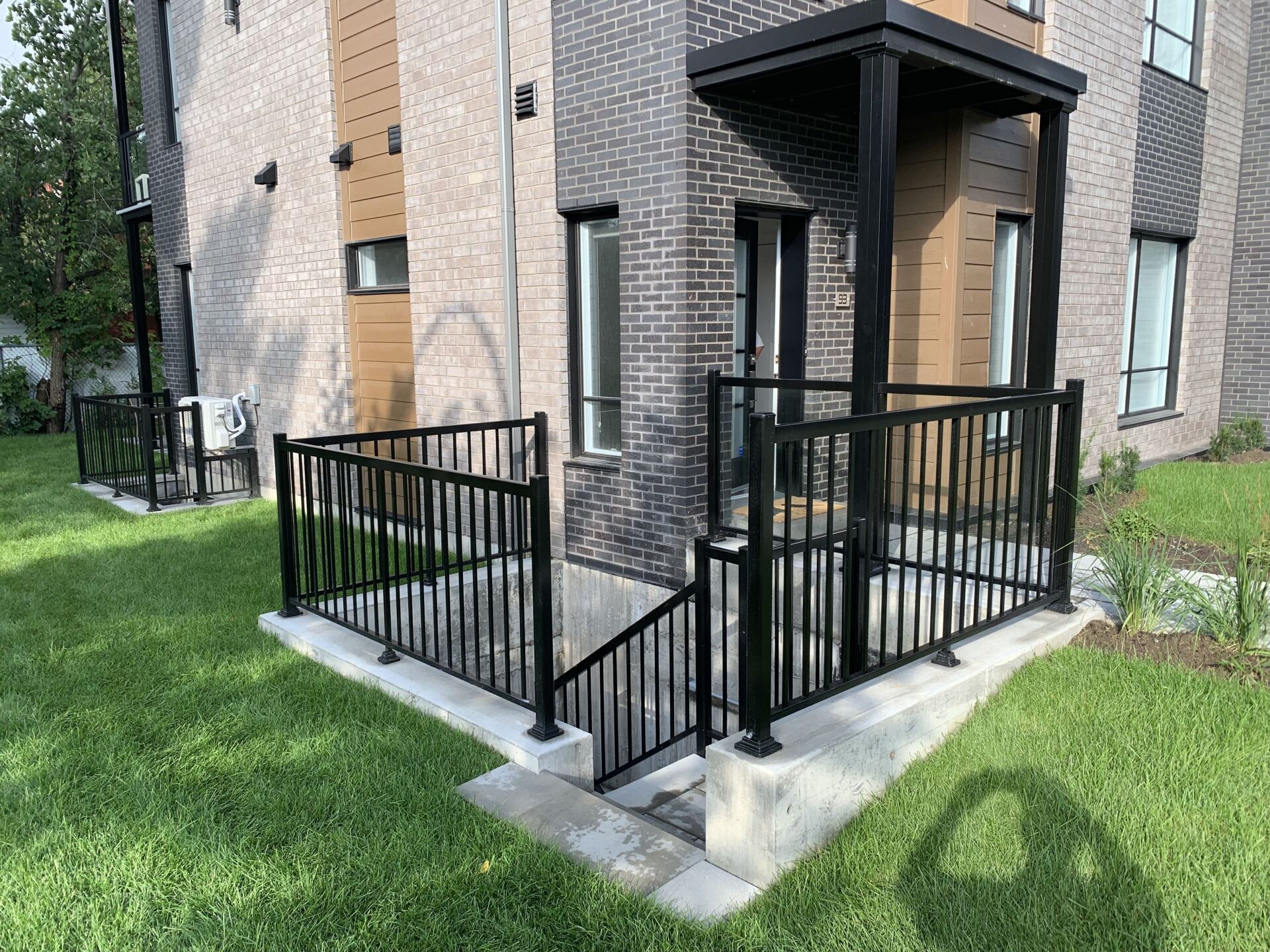 Une rambarde en métal noir entoure l'escalier du sous-sol et l'entrée d'un bâtiment moderne en briques. Une pelouse verdoyante entoure le tout.