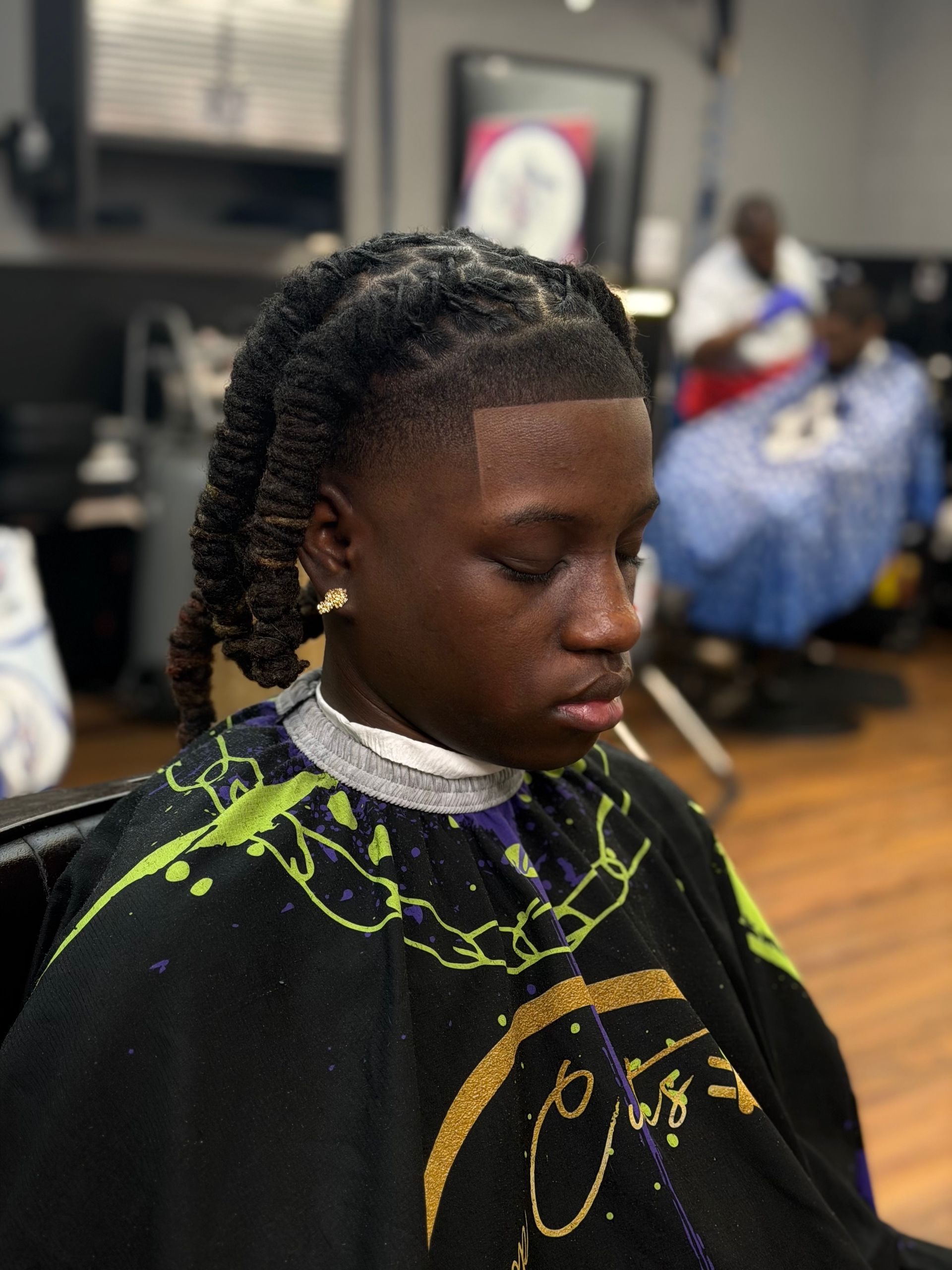 A person in a barber shop cape with a fade haircut and long, twisted hair, shown in profile.