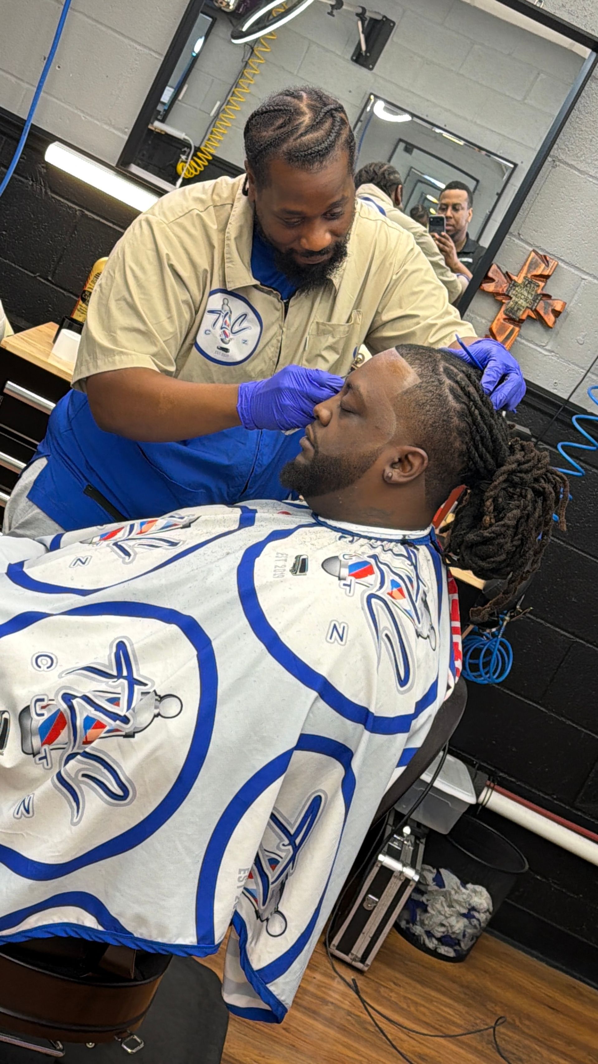 A barber in a tan shirt and blue gloves trims a client's hair in a salon with a reflective mirror in the background.