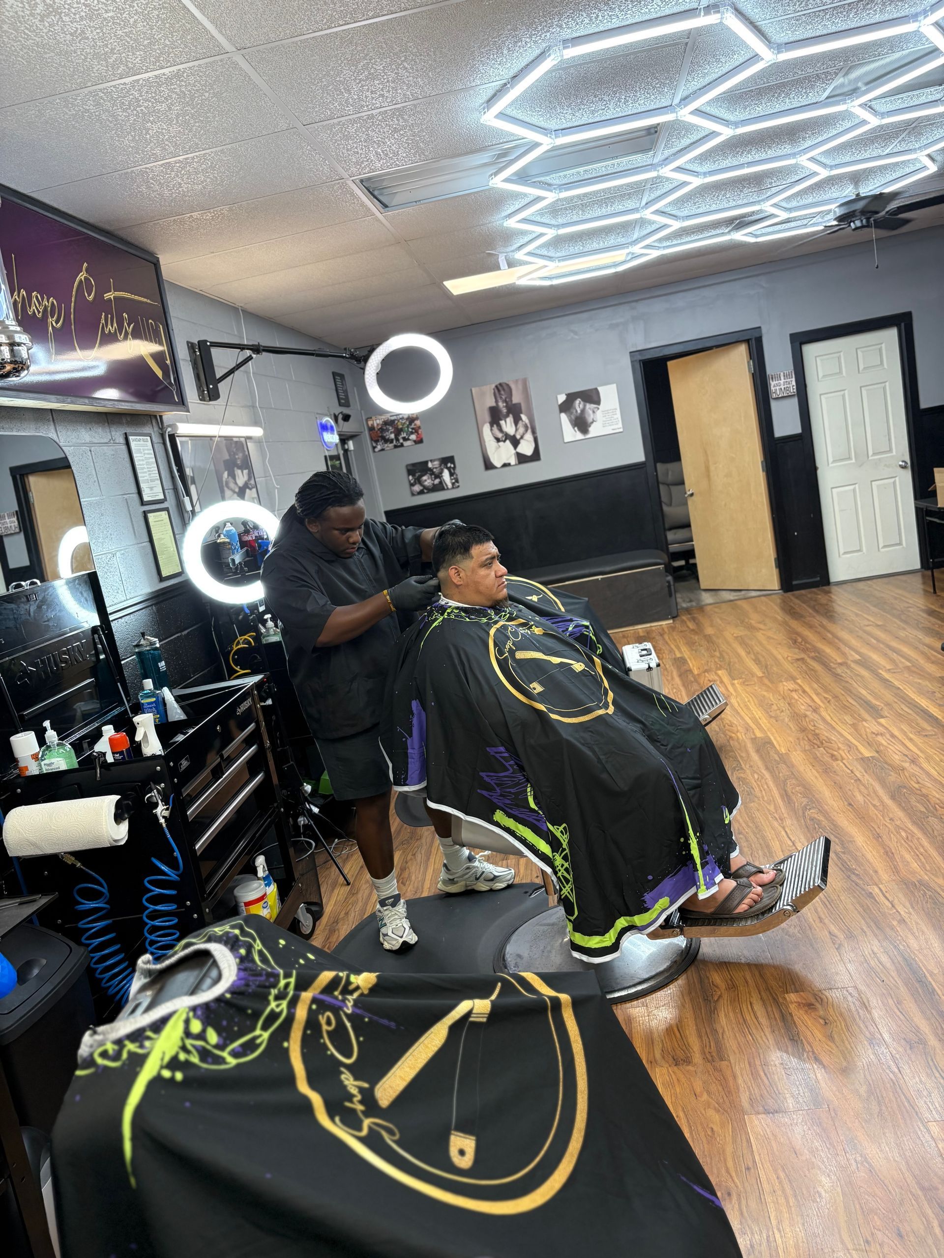 A barber cutting a client's hair in a modern salon with honeycomb ceiling lights, mirrors, and ring lights.