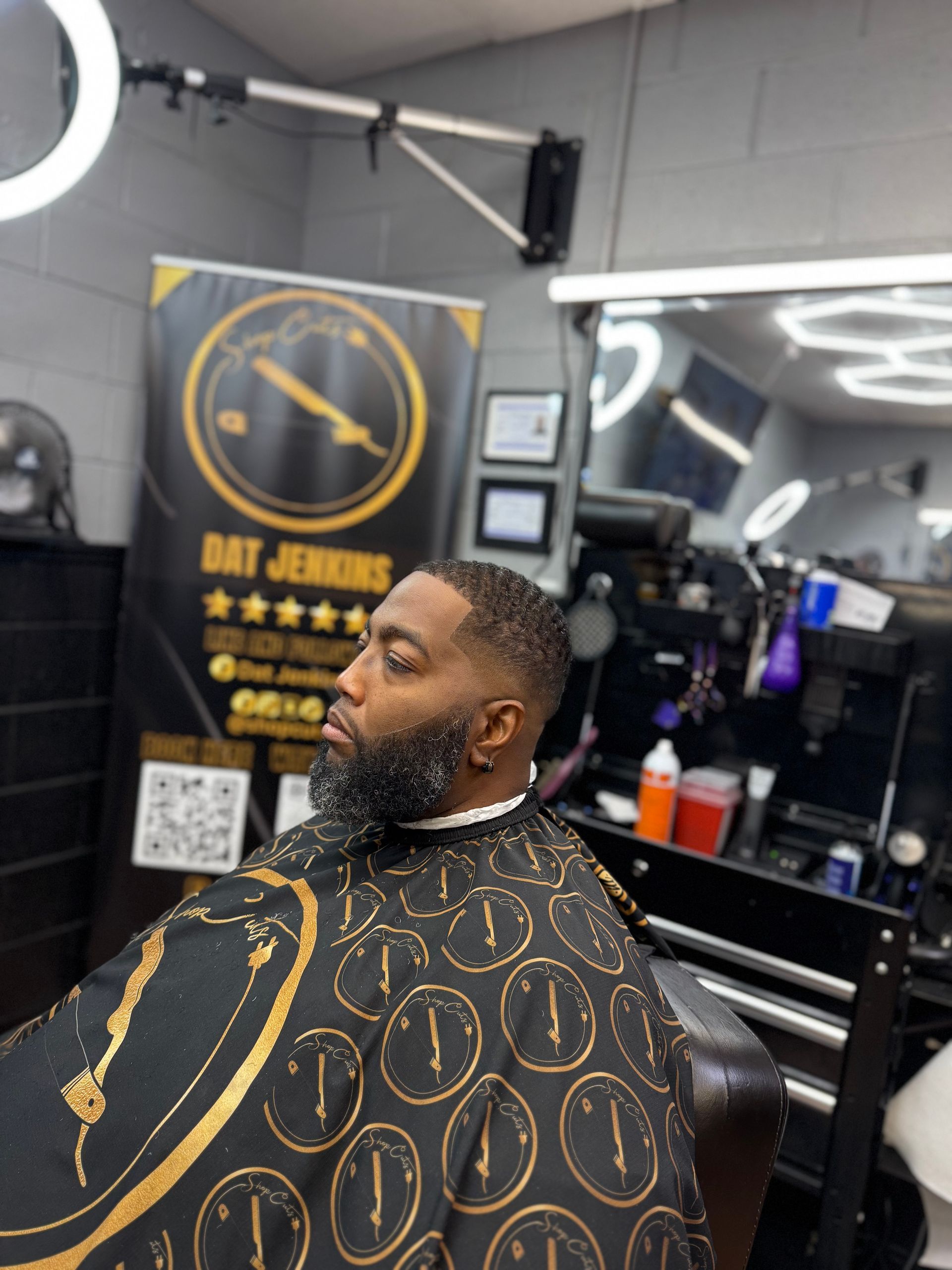 A person with a precise haircut and beard trim sits in a salon chair wearing a patterned cape in front of a barber sign.