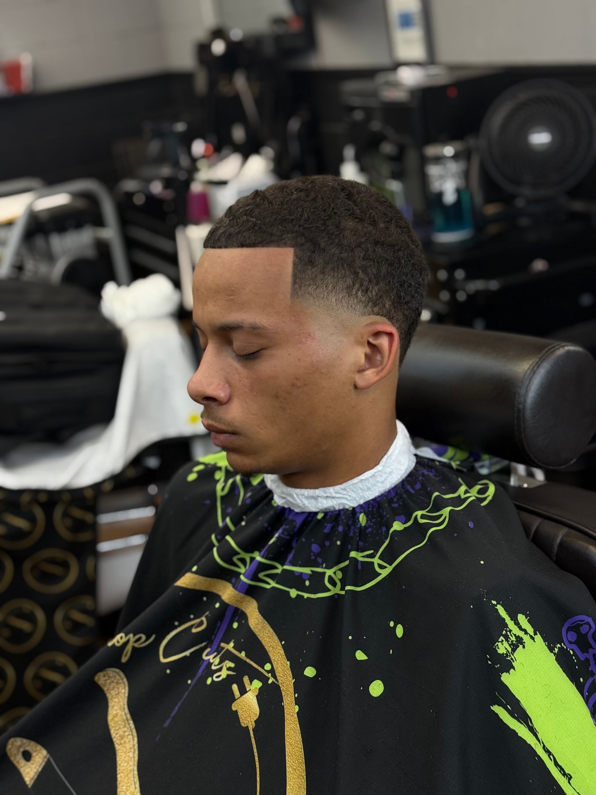 A person with a clean-cut fade haircut sits in a barber chair wearing a black and green cape in a barbershop.