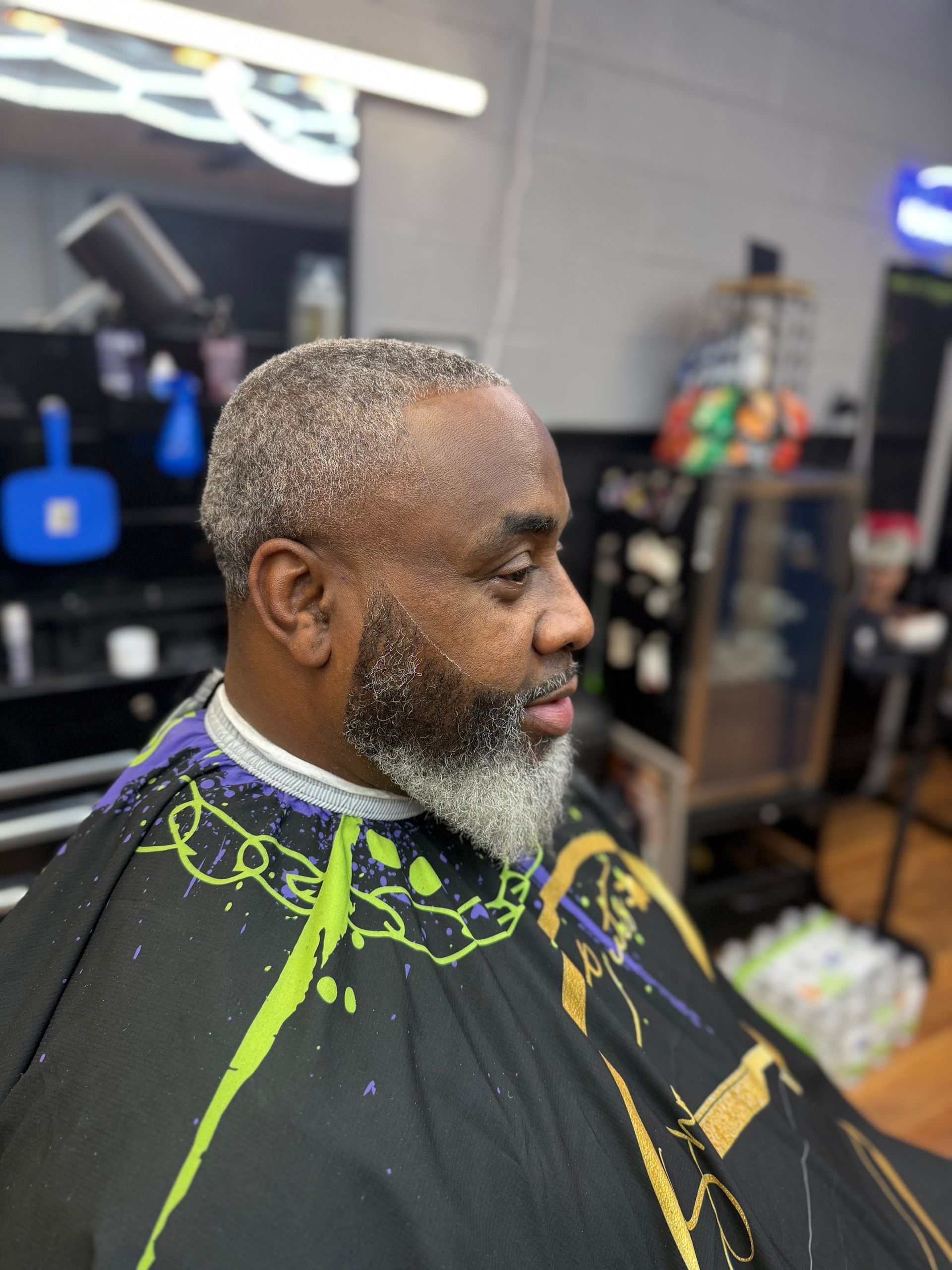 A side profile of a person in a barber’s cape showing a short, faded haircut and a neatly trimmed, gray-toned beard.