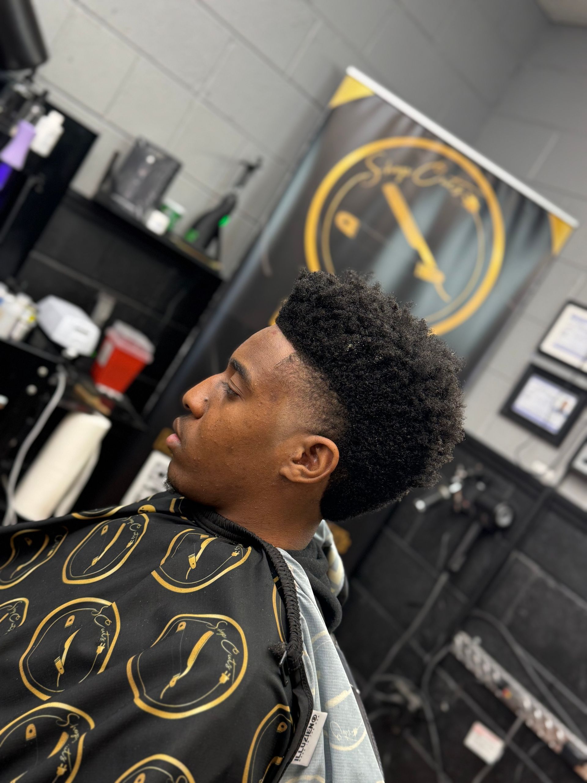 A side profile of a person with a textured fade haircut sitting in a barber chair wearing a black and gold cape.