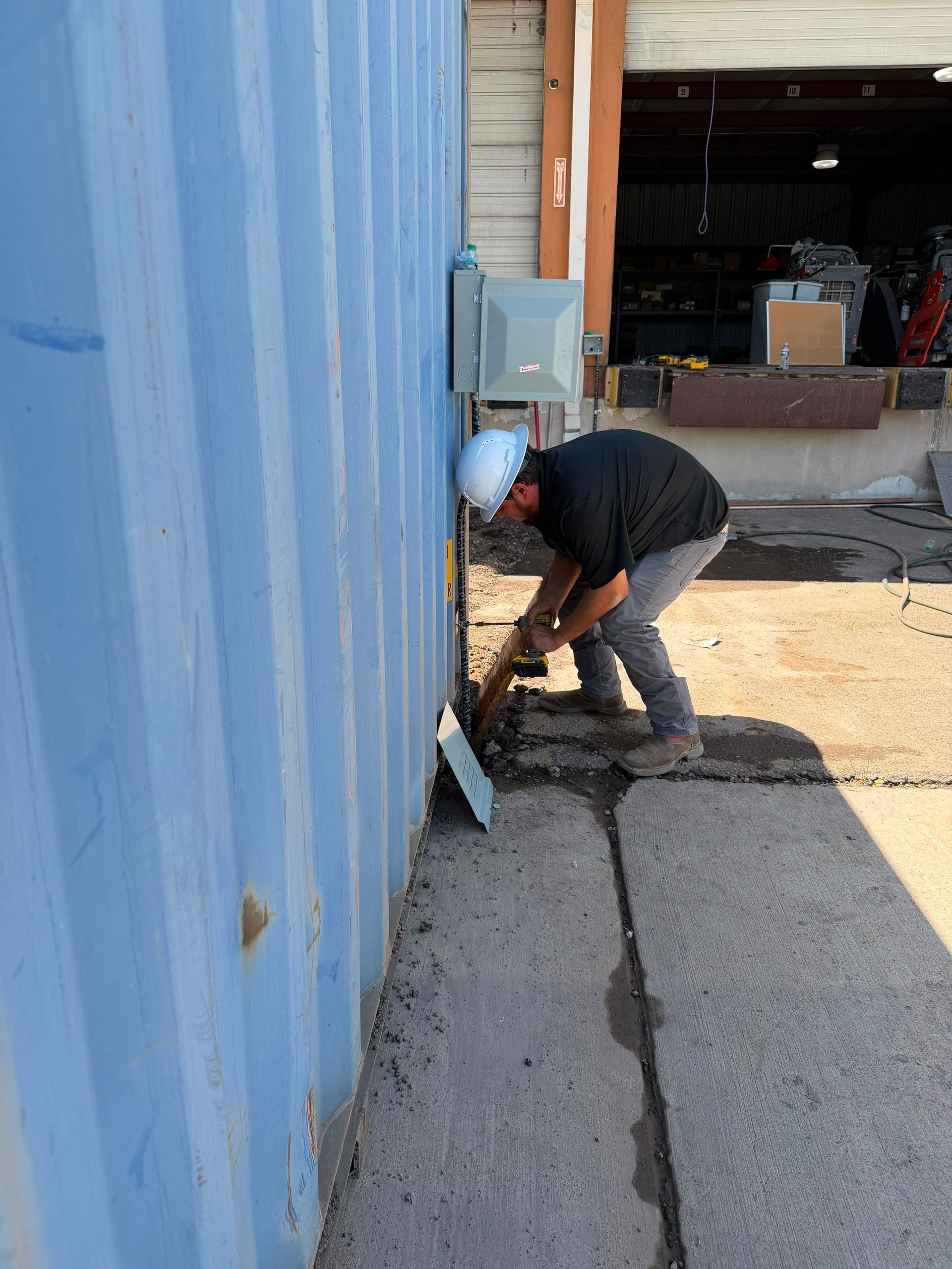 Worker bending beside a blue corrugated wall near an electrical box, outdoors by a garage opening.