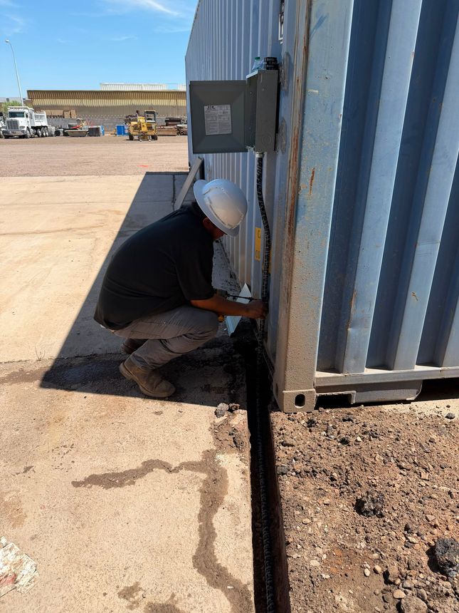 Worker crouched beside a corrugated metal structure, using tools near the base in an outdoor industrial site