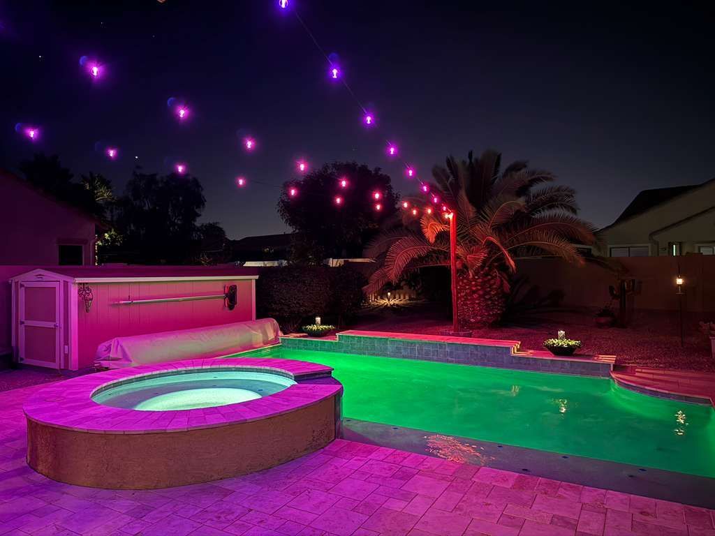 Nighttime backyard pool lit magenta and green, with glowing lanterns floating in the sky and palm trees nearby