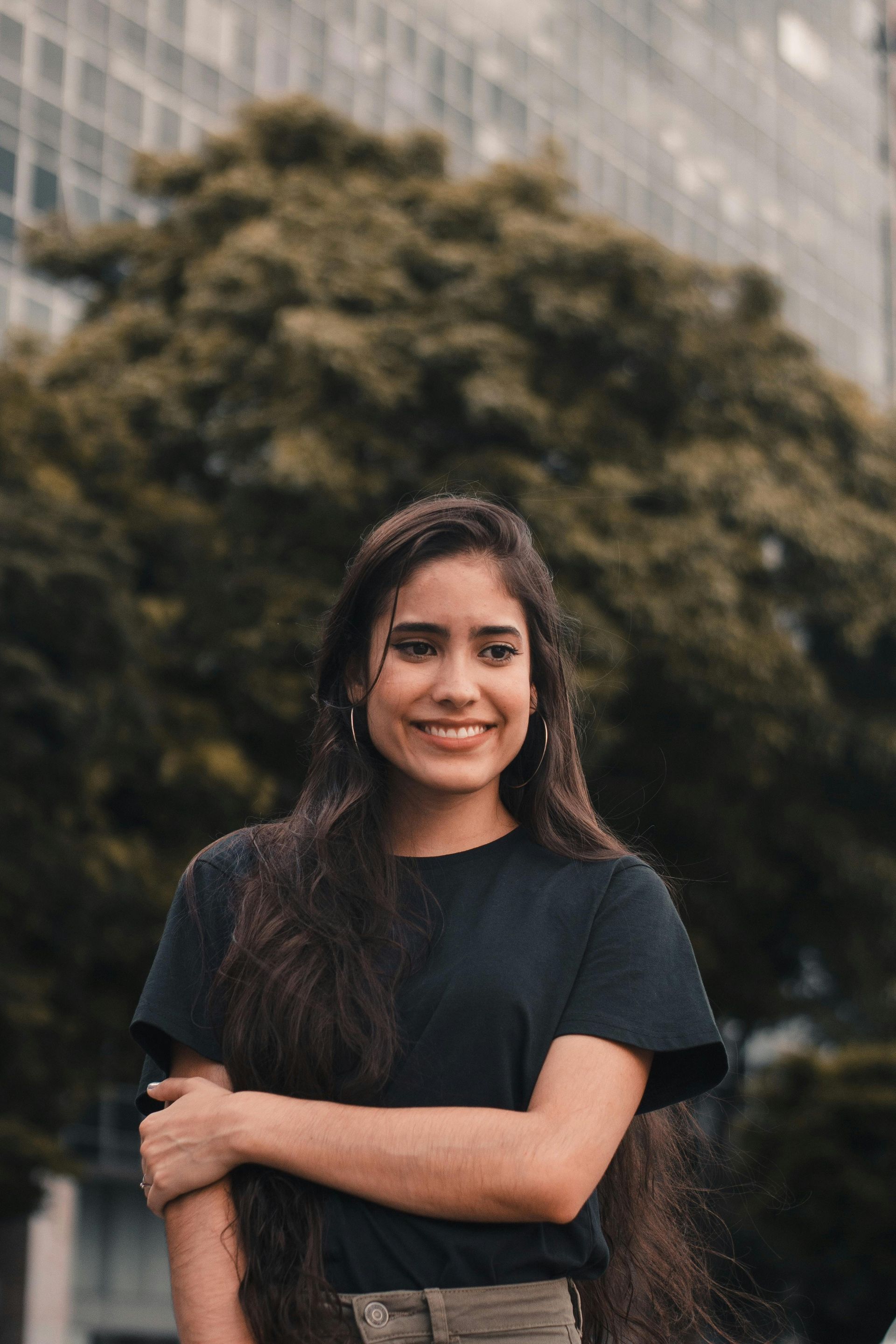 Woman standing outdoors with arms crossed, smiling in a black shirt against a blurred city backdrop