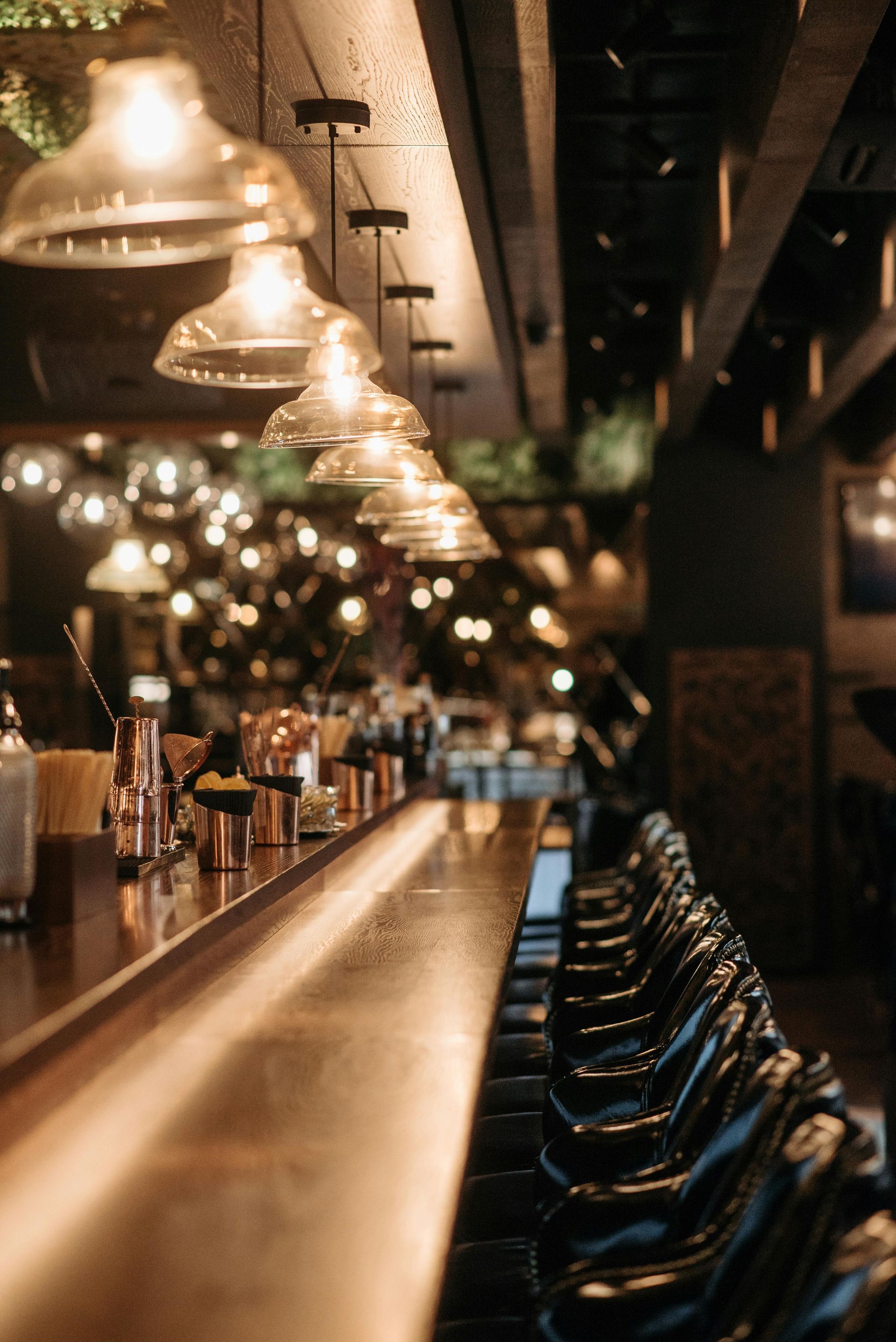 Dimly lit bar interior with hanging pendant lights, a wooden counter, and rows of stools