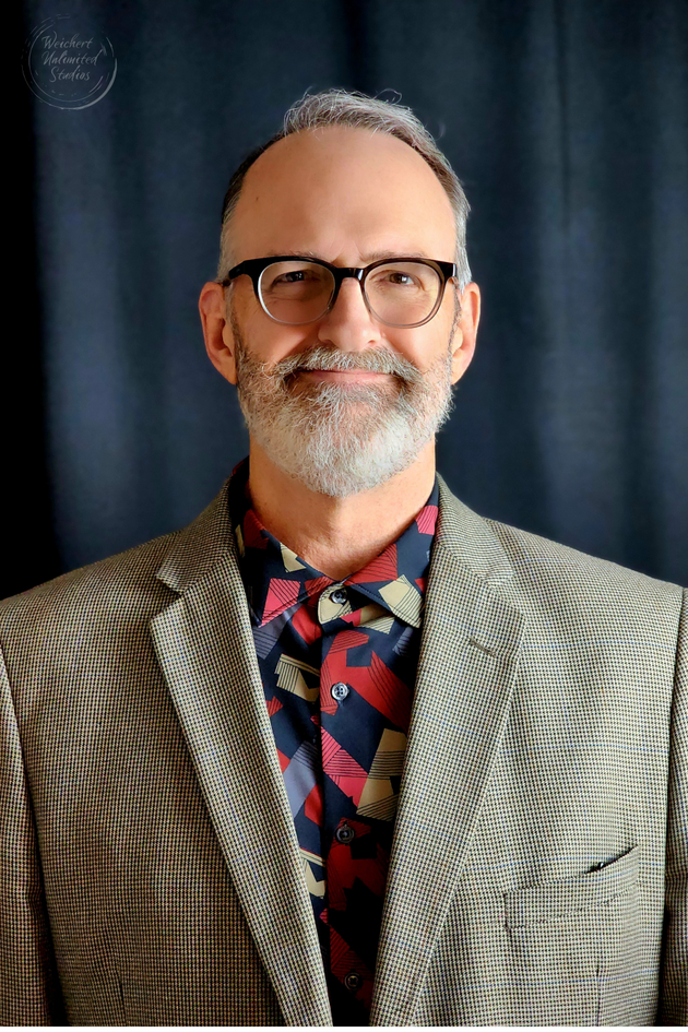 Portrait of a smiling man in glasses wearing a patterned blazer and floral tie against a dark backdrop