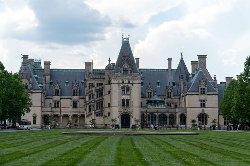 Historic stone mansion with ornate towers and arched windows, centered behind a wide green lawn under a cloudy sky