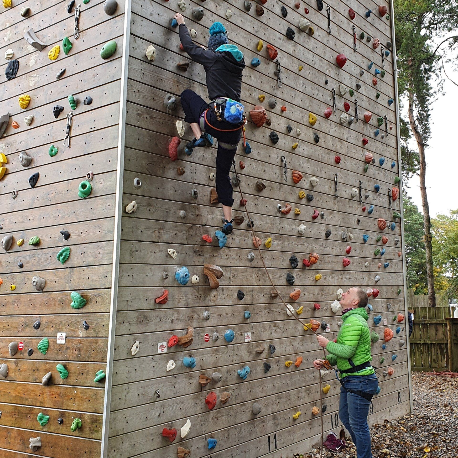 A man in a green jacket watches a woman climb a climbing wall