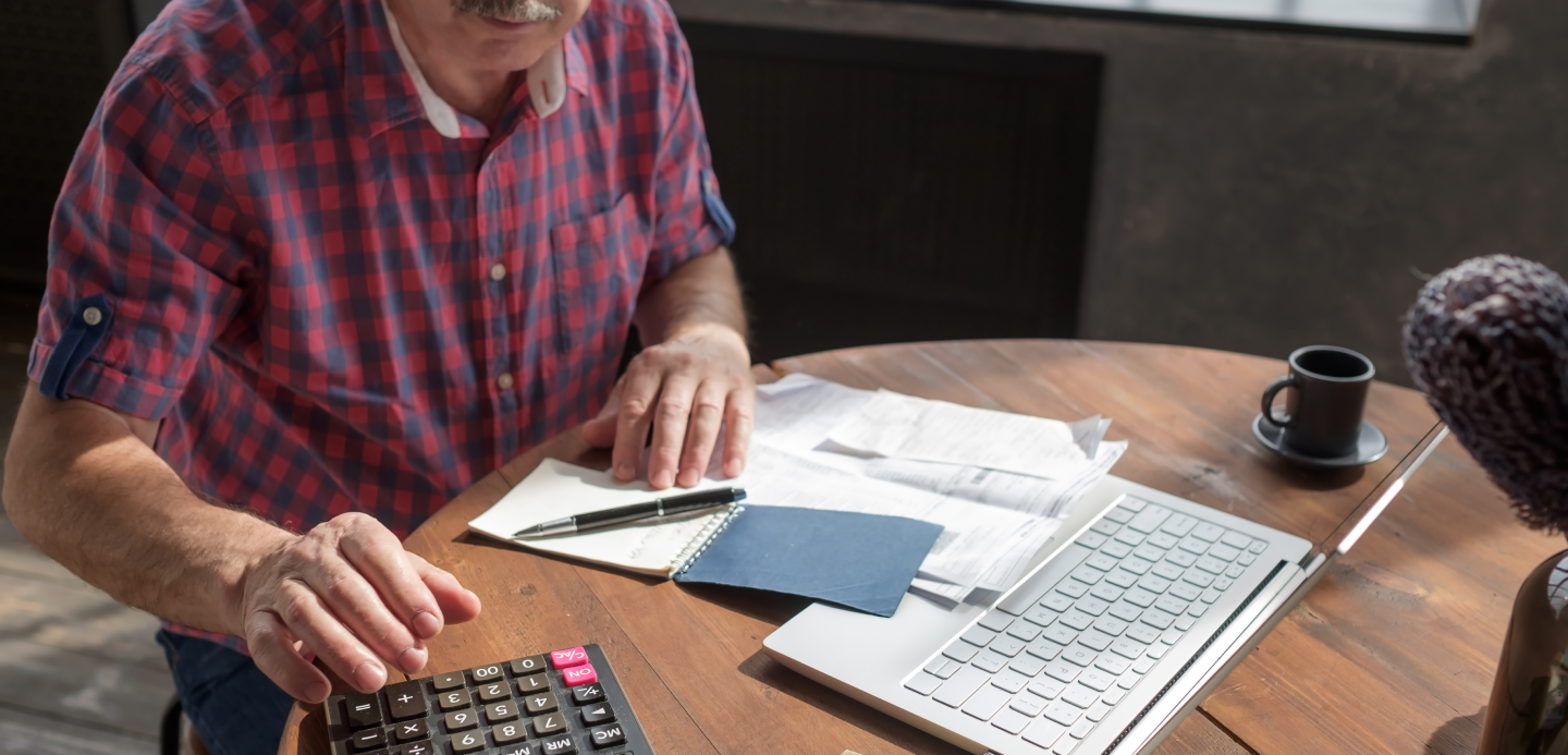 Um homem está sentado à mesa usando uma calculadora e um laptop.