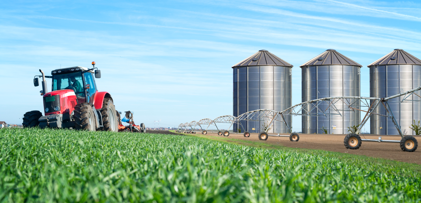 Um trator está arando um campo com silos ao fundo.