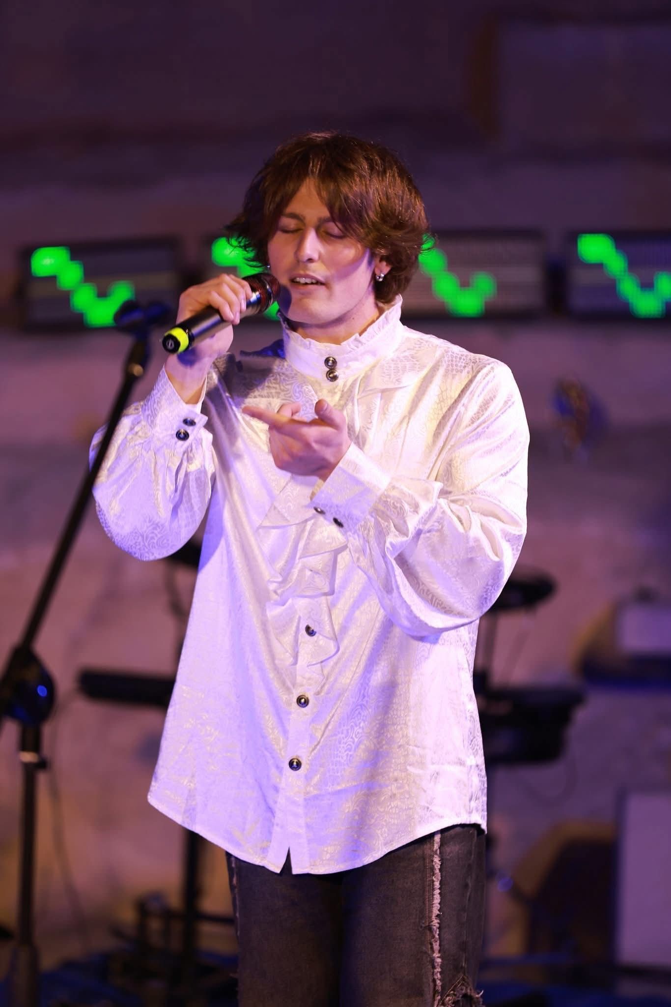 Un cantante con una camicia bianca tiene in mano un microfono sul palco, con luci verdi sullo sfondo.