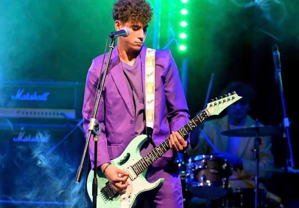 Un musicista con una giacca viola suona una chitarra elettrica bianca su un palco da concerto avvolto dal fumo.
