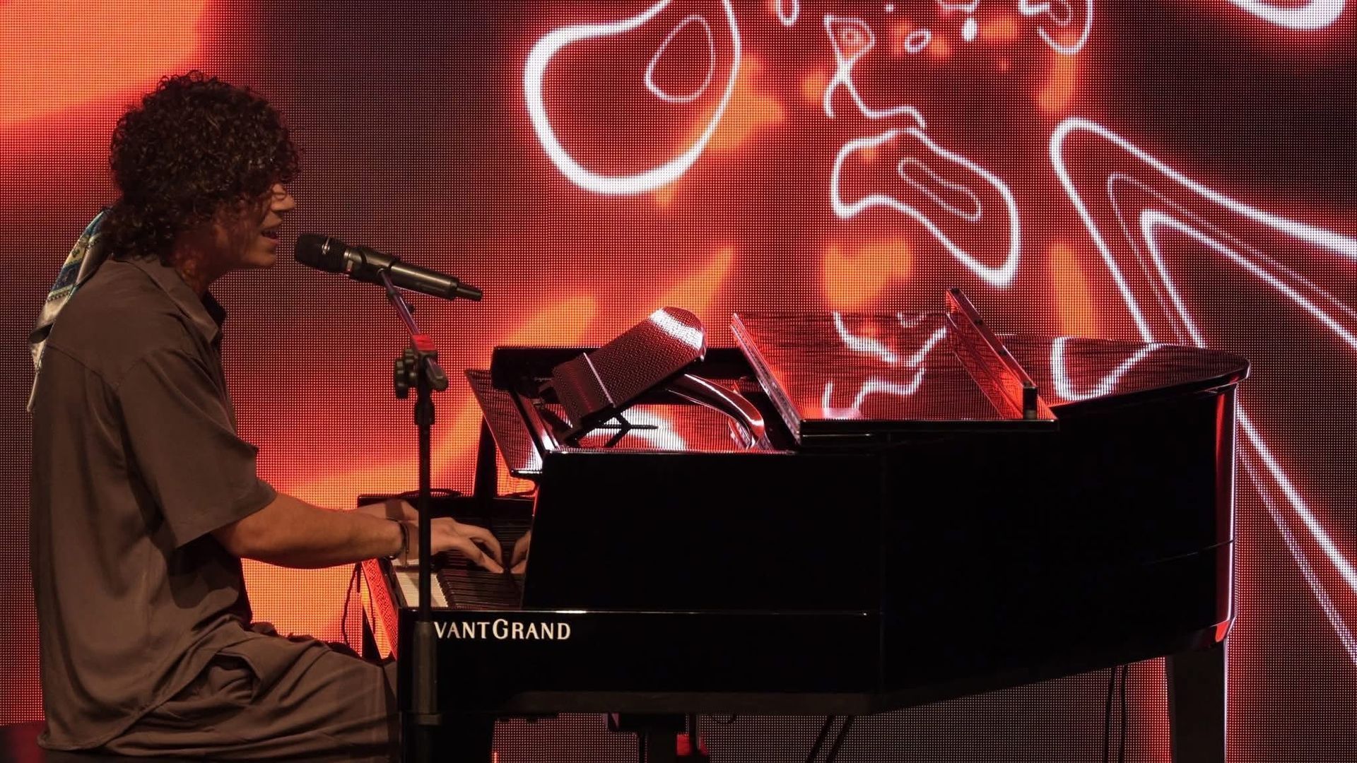 Un musicista suona un pianoforte a coda nero su un palco illuminato di rosso, con alle spalle un'opera d'arte astratta fatta di linee al neon bianche.