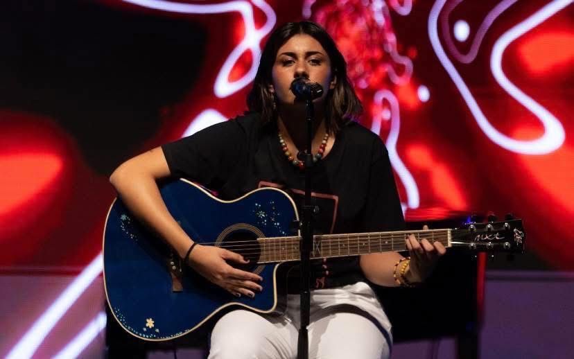Un musicista canta in un microfono mentre suona una chitarra acustica blu su un palco illuminato da luci rosse astratte.