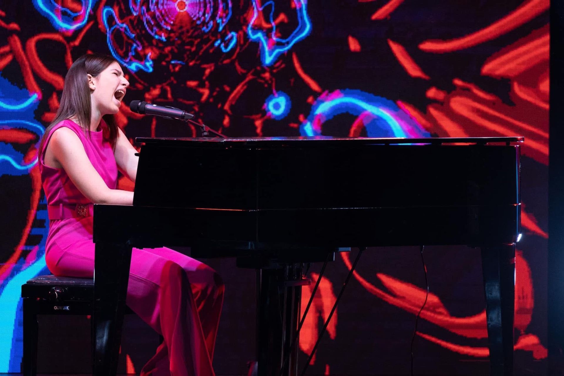 Una cantante in abito rosa si esibisce al pianoforte nero su un palco illuminato al neon.