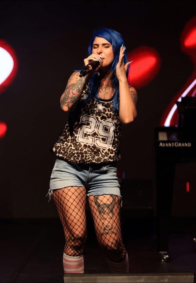 Una cantante dai capelli blu e tatuata si esibisce sul palco, tenendo in mano un microfono sotto luci rosse.